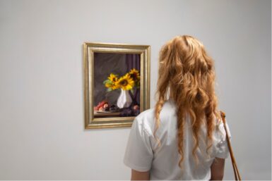 Femme aux cheveux bouclés et avec un tee-shirt blanc observant un tableau qui représente des tournesols dans un vase.